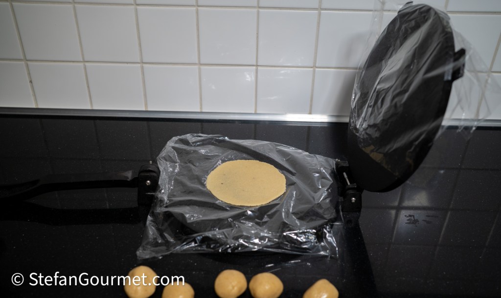 A tortilla press with a flattened circle of dough in between two sheets of plastic wrap, surrounded by balls of dough on a black countertop.