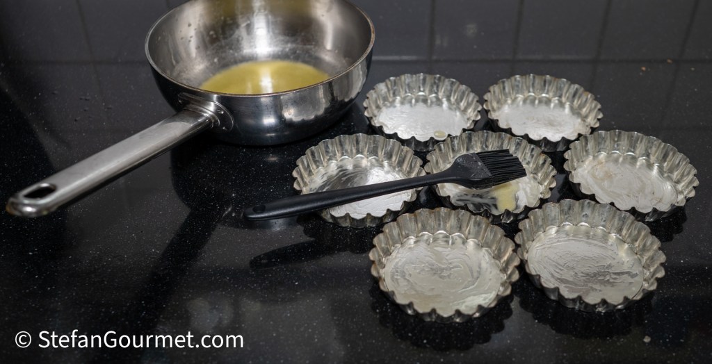 A stainless steel saucepan with melted butter sits next to eight greased tartlet tins on a black surface, with a pastry brush placed on one of the tins.
