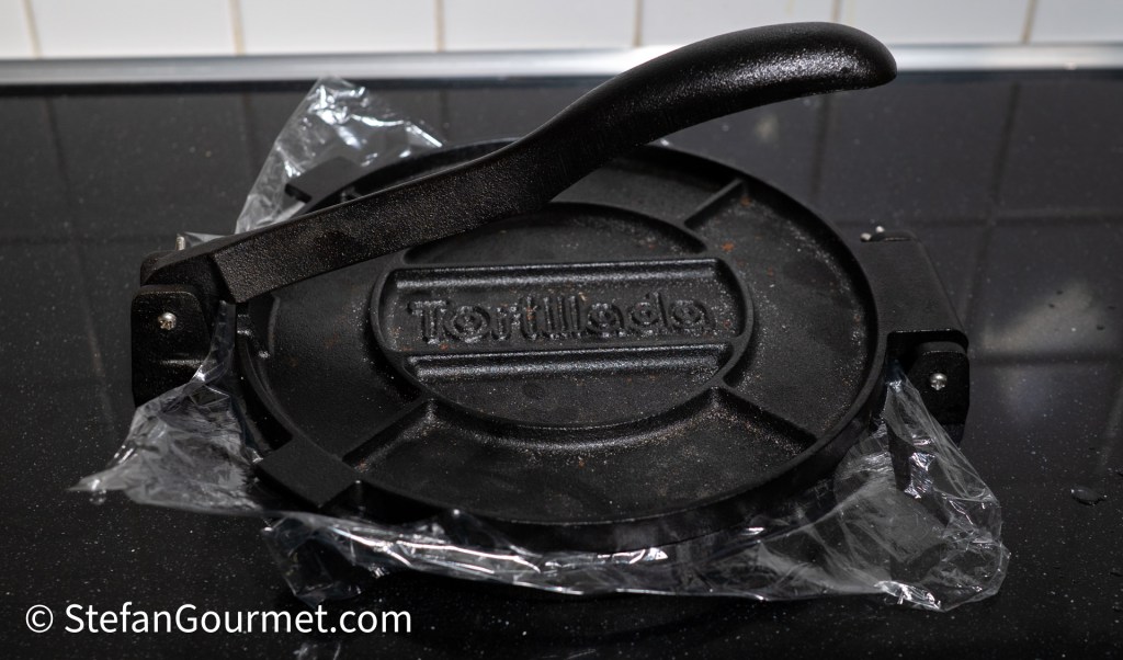 A tortilla press placed on a black countertop, covered with plastic wrap for food preparation.