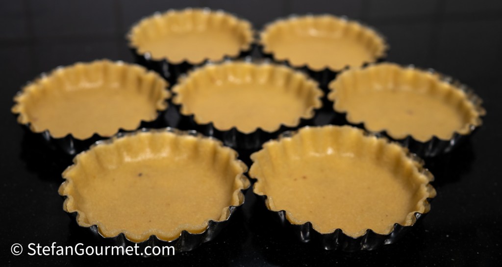 Eight almond tart shells waiting to be filled, set against a dark countertop.