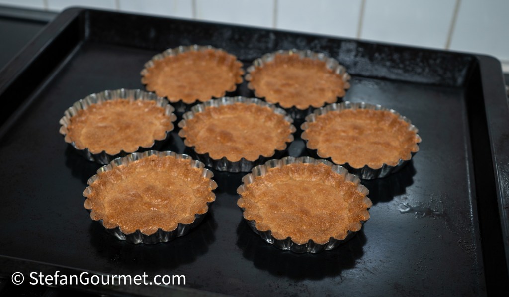 Baked almond tart shells cooling on a black baking tray.