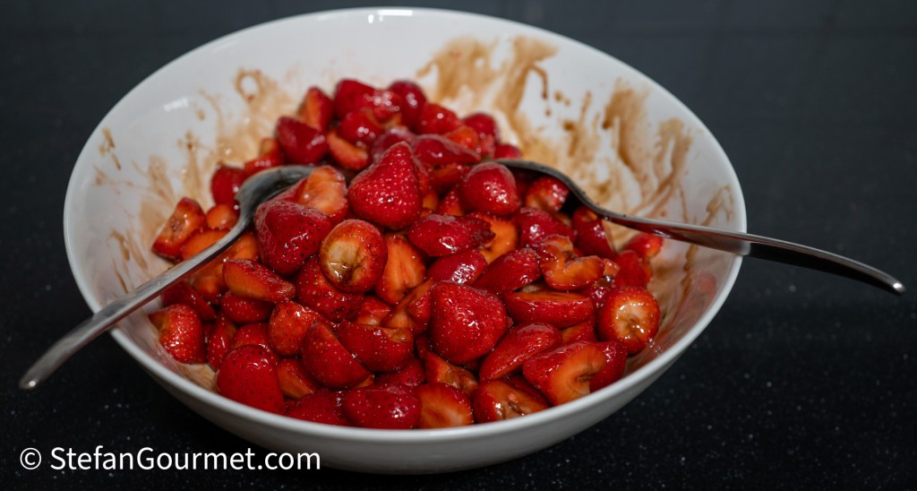 A white bowl filled with halved strawberries coated in honey and balsamic vinegar, with two serving spoons resting on the edge.