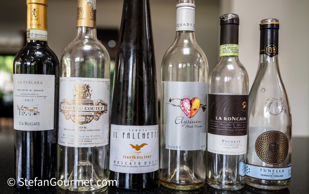 A collection of emptied wine bottles on a dark countertop, showcasing various labels including La Perlora, Château Coutet, Tenuta Il Falchetto, Elysium, La Roncaia, and Tunella.