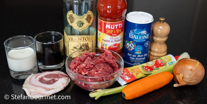 Ingredients for Bolognese sauce arranged on a black countertop, including ground beef, pancetta, vegetables, wine, tomato products, and seasonings.