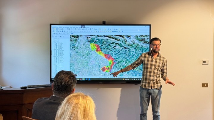 A presenter stands in front of a large screen displaying a colorful map while explaining data related to vineyards.