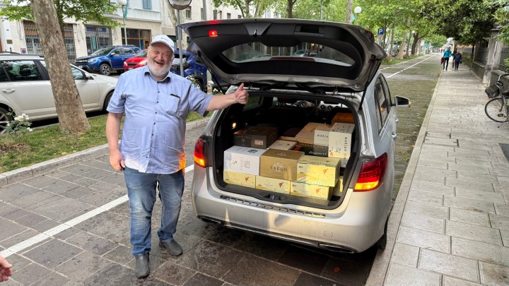 A smiling man stands beside a car with its trunk open, showcasing numerous boxes of wine being transported after a wine tour.