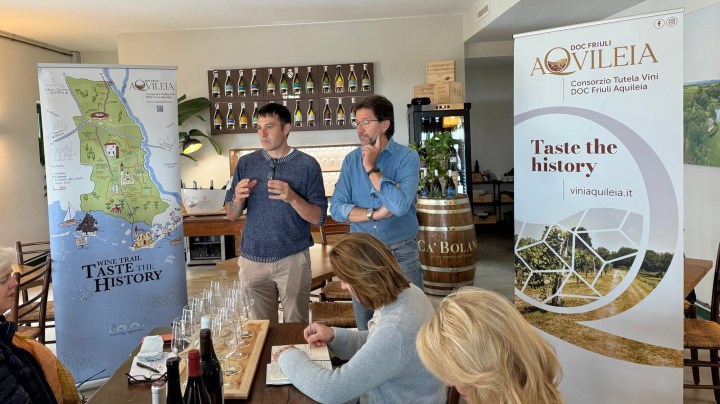 Two presenters discussing wine in a tasting room with a map and banner for DOC Friuli Aquileia in the background, while participants listen attentively.