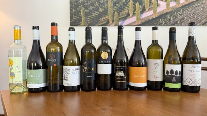 A selection of white wines from various wineries in Friuli Aquileia DOC displayed on a wooden table, with a vineyard landscape in the background.
