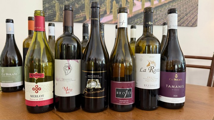 A selection of wine bottles displayed on a wooden table, featuring various labels and types, including Pinot Bianco and Refosco from the Friuli Aquileia region.