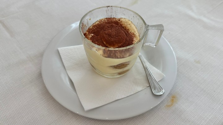 A glass dish of tiramisù topped with cocoa powder, served on a white plate with a napkin.