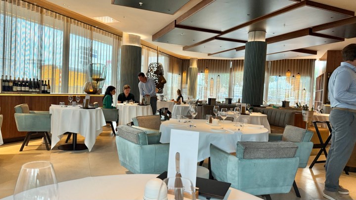 A dining room in a modern restaurant featuring elegant tables set for diners, with stylish decor and large windows allowing natural light. Waitstaff is attending to guests while others enjoy their meals.