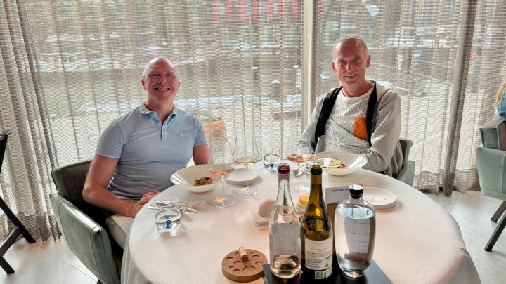 Two men seated at a dining table in a restaurant, smiling and enjoying a meal, with plates of food and wine bottles visible.