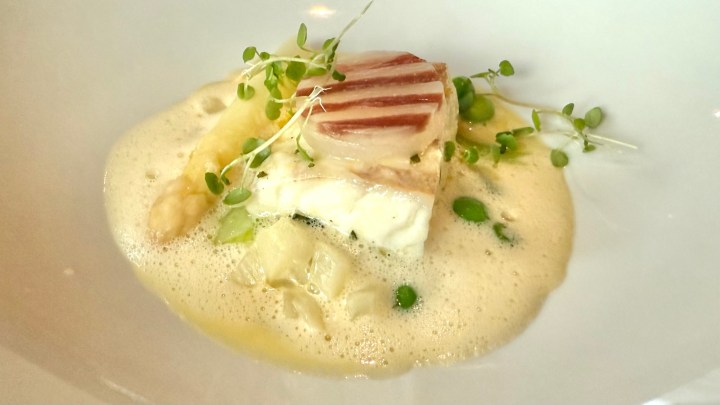 A beautifully plated dish featuring a piece of cooked fish, possibly sea bass, on a bed of creamy sauce with green peas and microgreens garnishing the top.