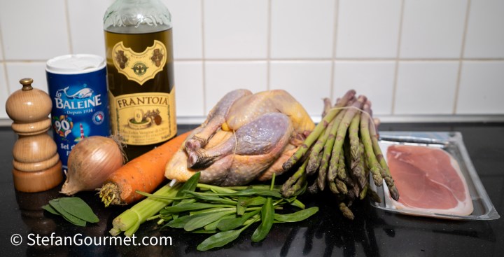 An assortment of ingredients for a dish, including a guinea fowl, fresh asparagus, sea lavender, a carrot, an onion, a bottle of olive oil, and a package of prosciutto, arranged on a kitchen countertop.