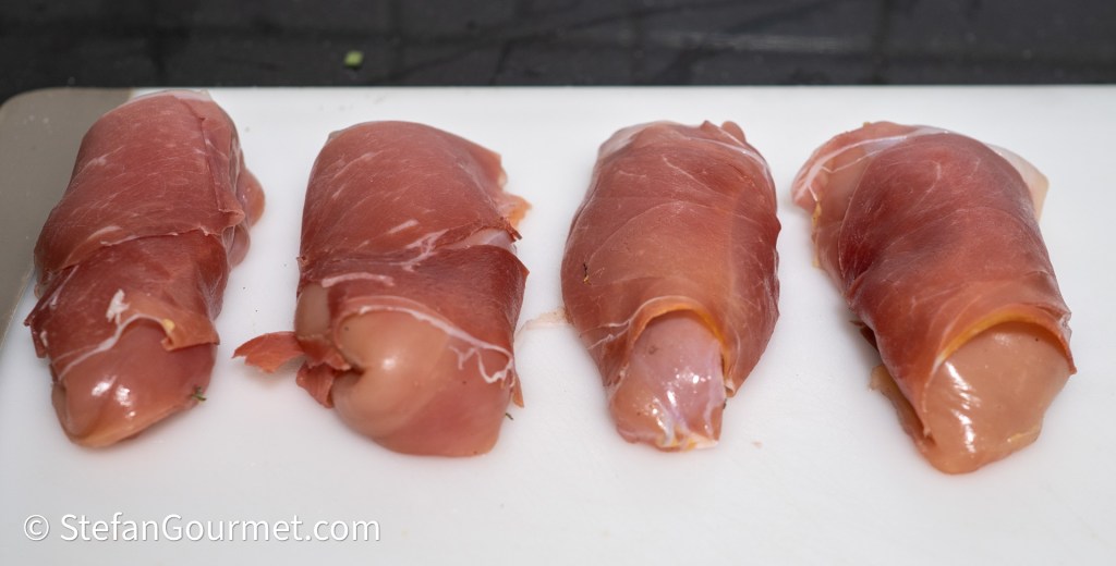Four rolled guinea fowl breast fillets wrapped in prosciutto, resting on a cutting board.