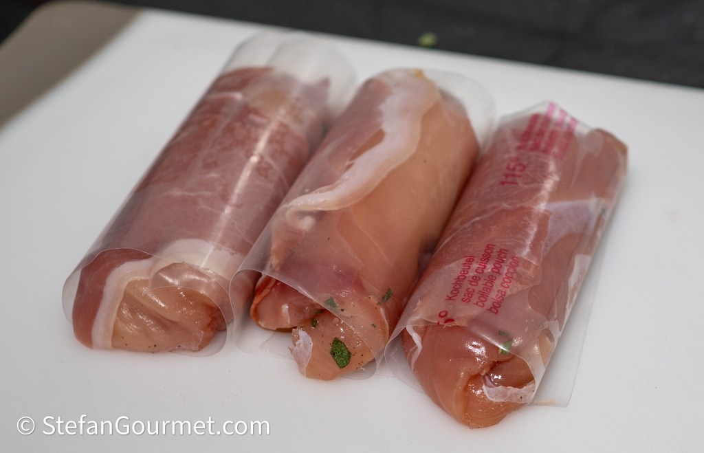 Three rolled guinea fowl breast fillets wrapped in prosciutto and sealed in vacuum bags, resting on a cutting board.