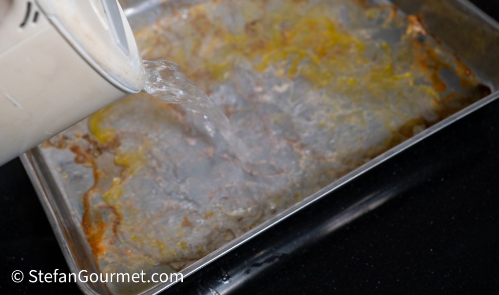 Pouring boiling water into a metal roasting tray to deglaze and collect drippings.