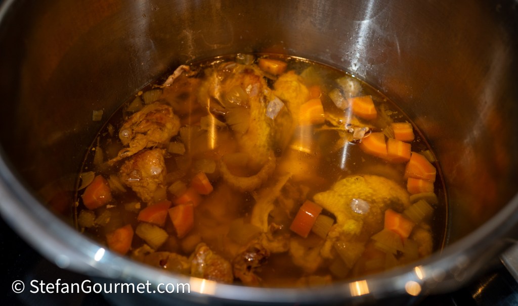 A pot containing roasted guinea fowl pieces simmering with chopped vegetables like carrots, onion, and celery in a flavorful broth.