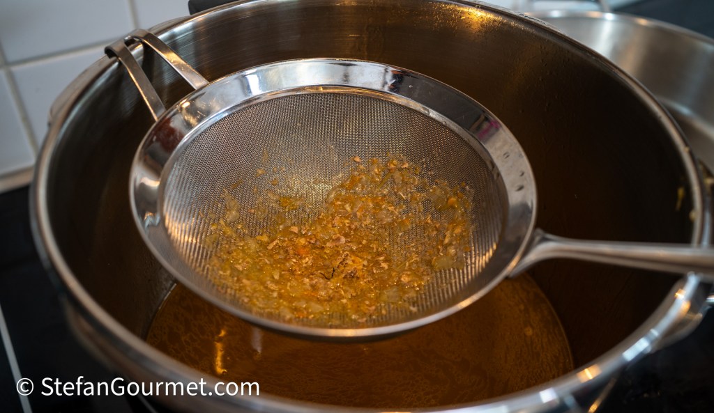 A fine mesh strainer sitting above a pot filled with a golden-brown liquid, filtering out solids from a stock or broth.