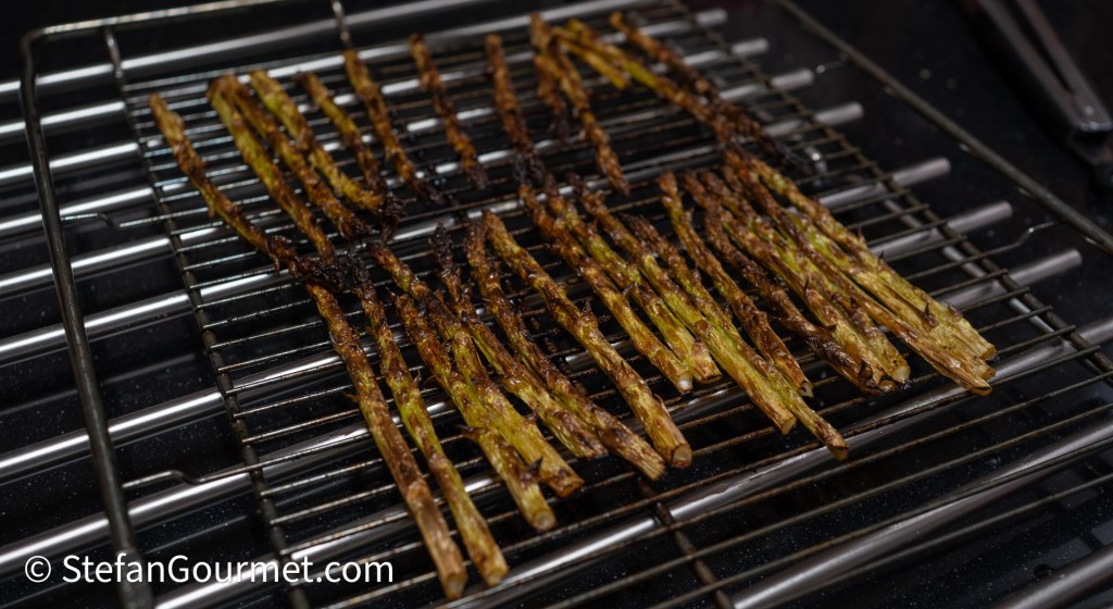 A tray of roasted asparagus with a slightly charred appearance, arranged on a wire rack, showing various shades of green and brown.