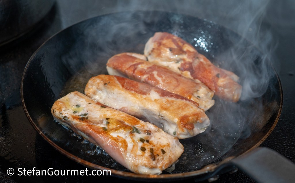 Grilled guinea fowl breast rolls wrapped in prosciutto, cooking in a frying pan with steam rising.