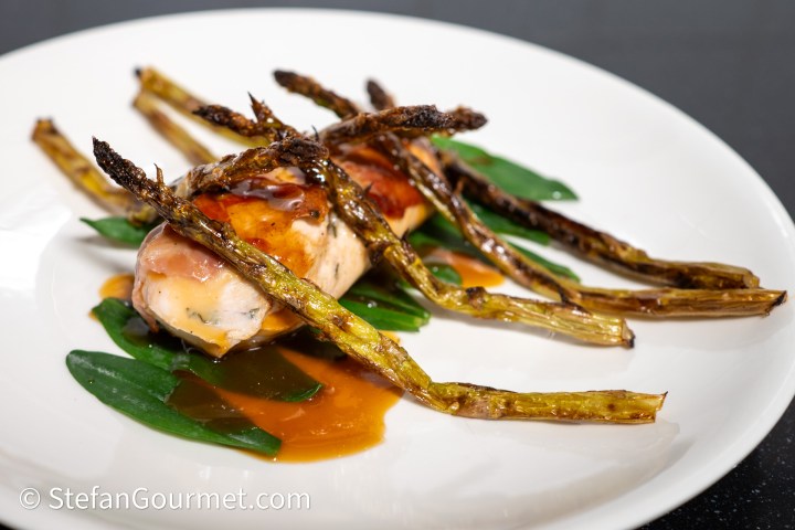 A plated dish featuring rolled guinea fowl wrapped in prosciutto, served on a bed of green leaves, topped with roasted asparagus and a drizzle of jus.