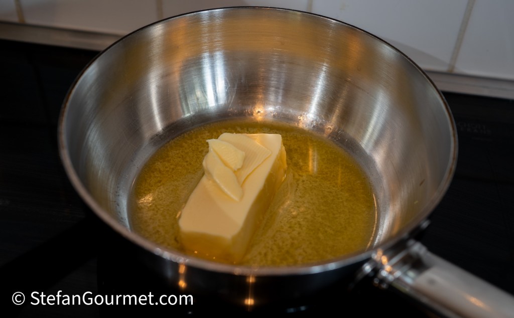 A saucepan on a stovetop containing a block of butter melting in a pool of liquid butter.