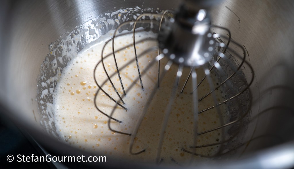 A close-up view of a metal mixing bowl with foamy egg and sugar mixture, fitted with a whisk attachment.