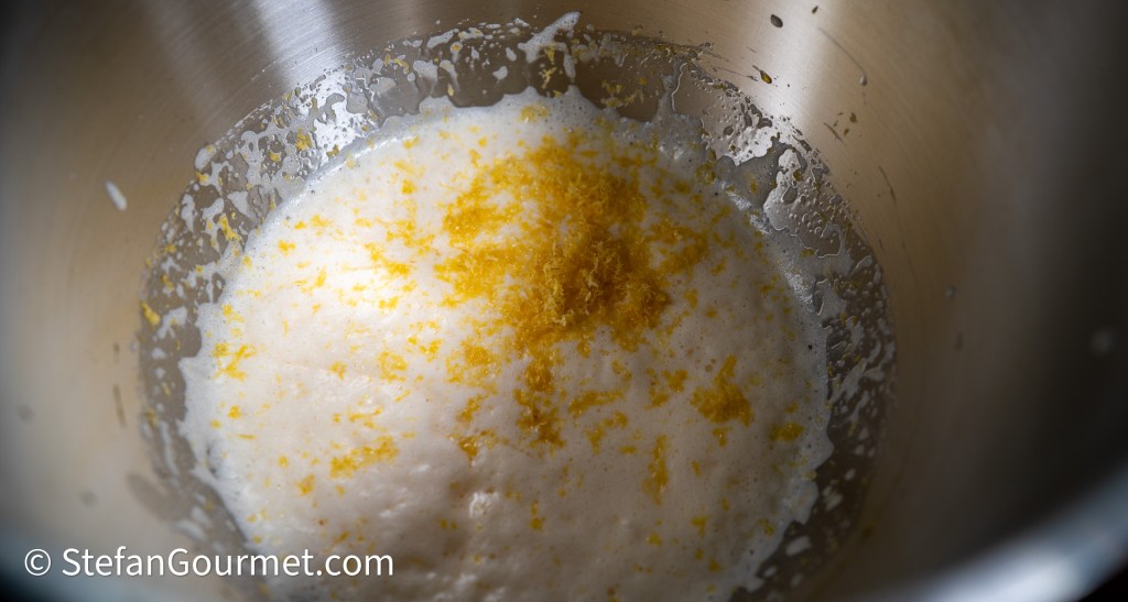 A stainless steel mixing bowl containing foamy whipped eggs and sugar, topped with grated lemon zest.
