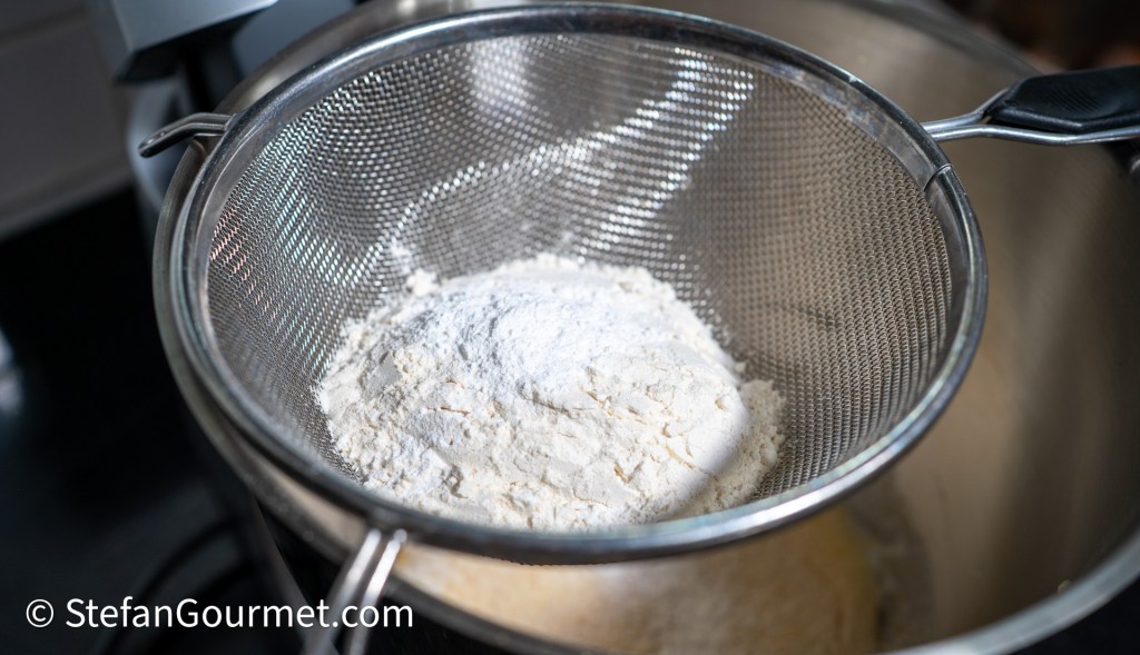 A close-up view of a fine sieve containing sifted flour, with the background showing a mixing bowl and kitchen equipment, capturing the preparation process for baking.