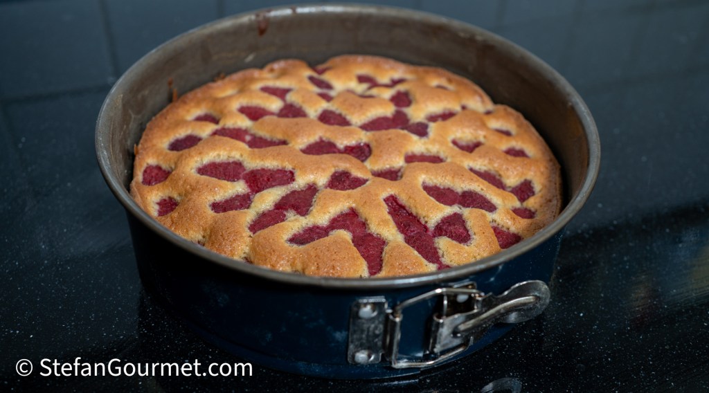 Baked raspberry and almond cake in a round springform pan, showing a golden-brown crust with visible raspberry swirl patterns on top.