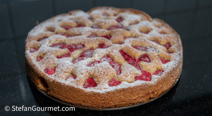 A raspberry and almond cake dusted with icing sugar, featuring a golden brown crust and visible pieces of fresh raspberries on top, placed on a dark marble countertop.