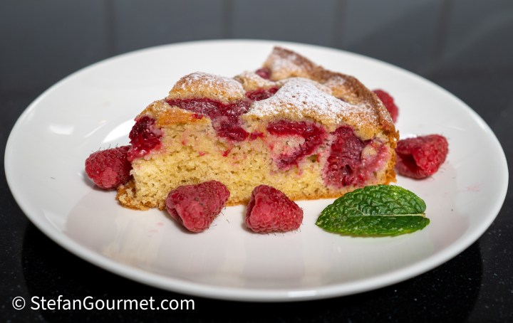 A slice of raspberry and almond cake served on a white plate, garnished with fresh raspberries and a sprig of mint.