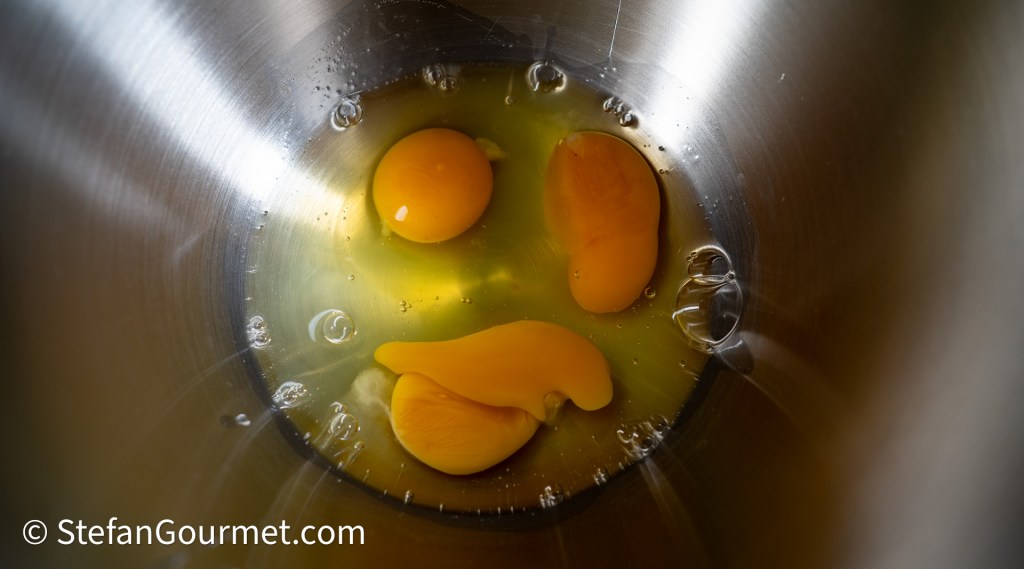 Three egg yolks and egg whites in a stainless steel mixing bowl.