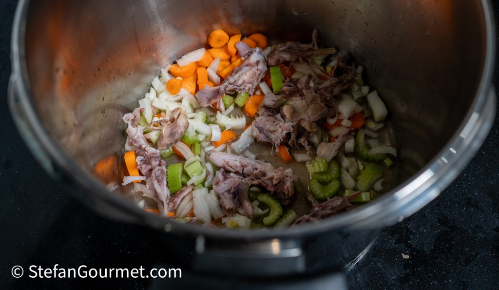 A silver pot filled with chopped vegetables including carrots, celery, and onion, along with pieces of rabbit meat.