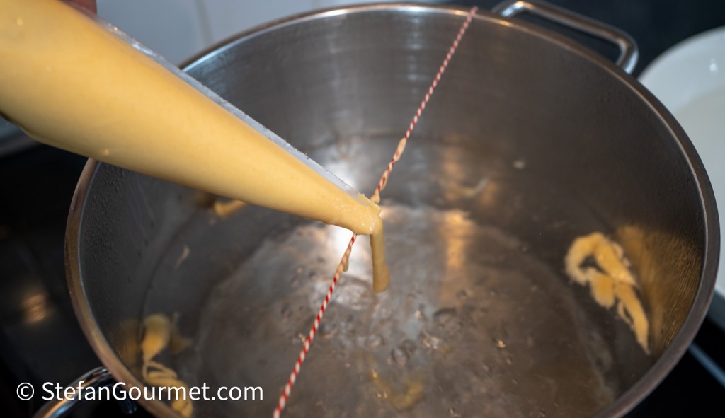 A piping bag filled with pasta batter is positioned above a pot of boiling water, with a piece of kitchen twine tied across the pot's edges to guide the dripping pasta.