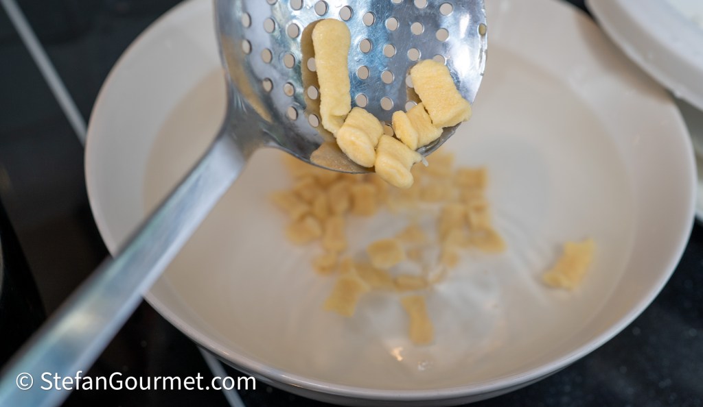 A slotted spoon lifting freshly cooked girini pasta from a pot, with some pieces placed in a white dish.