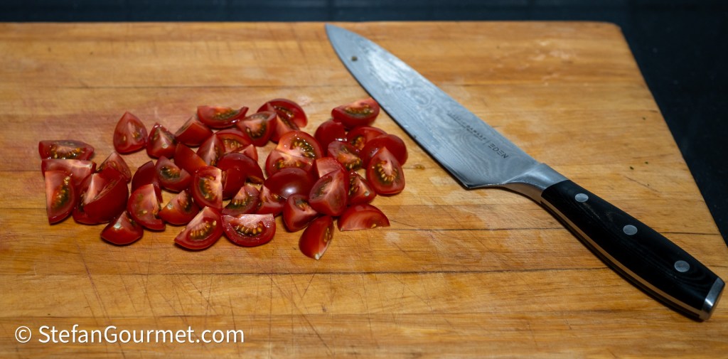 Sliced cherry tomatoes arranged on a wooden cutting board next to a sharp kitchen knife.
