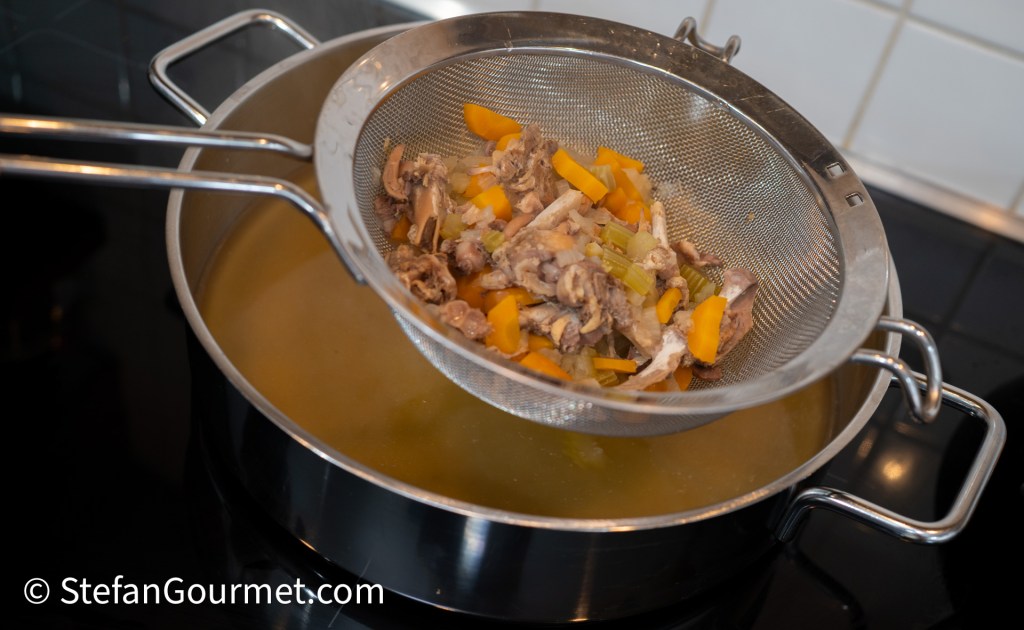 Strainer holding cooked rabbit bones and vegetables over a pot of golden broth.