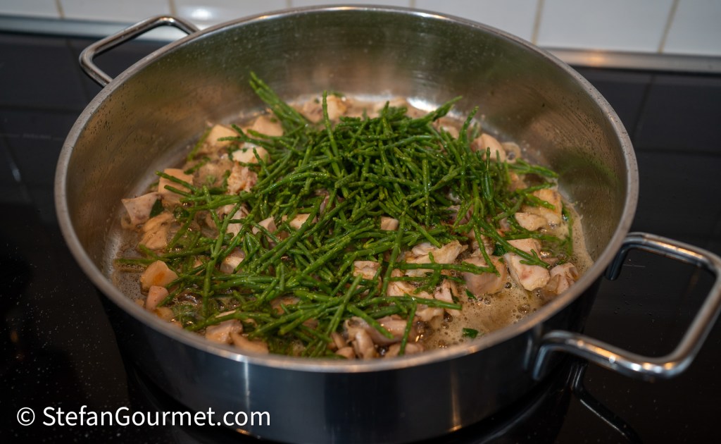 A pot on a stovetop containing rabbit meat and fresh samphire cooking together in a flavorful broth.