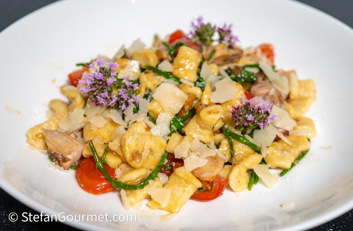 A plated dish featuring _girini_ pasta mixed with cherry tomatoes, rabbit meat, and samphire, garnished with fresh oregano flowers and Parmesan cheese.