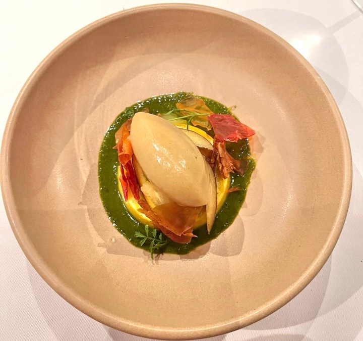 A dessert plate featuring a smooth sorbet atop a yellow pastry base, garnished with colorful dried leaves and surrounded by a green sauce.