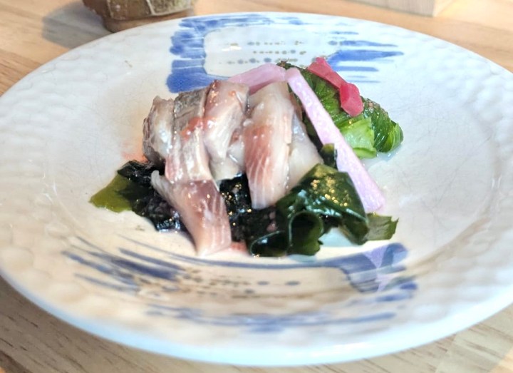 A plate featuring slices of fish on a bed of seaweed, garnished with pink pickled vegetables and green leafy elements, showcasing a delicate and colorful presentation.