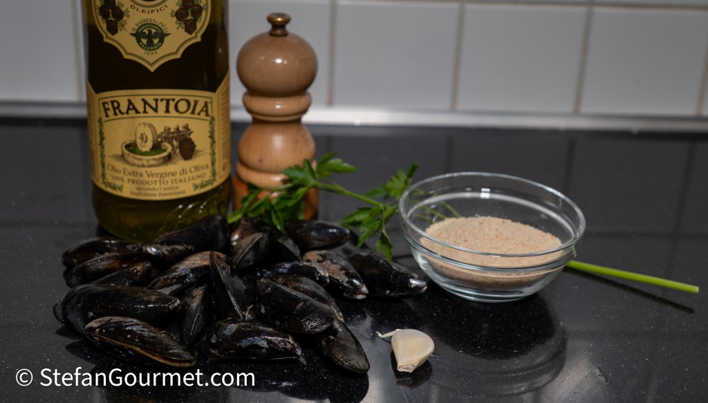 Ingredients for gratinated mussels, including mussels, garlic, parsley, olive oil, and breadcrumbs on a kitchen countertop.