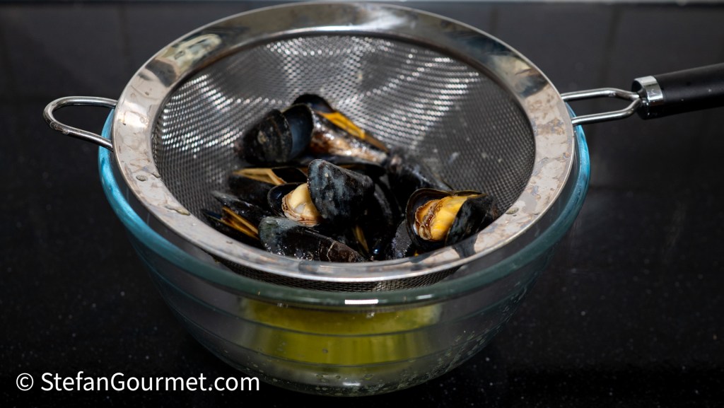 A colander filled with freshly cooked mussels resting over a bowl, with some juices visible at the bottom.