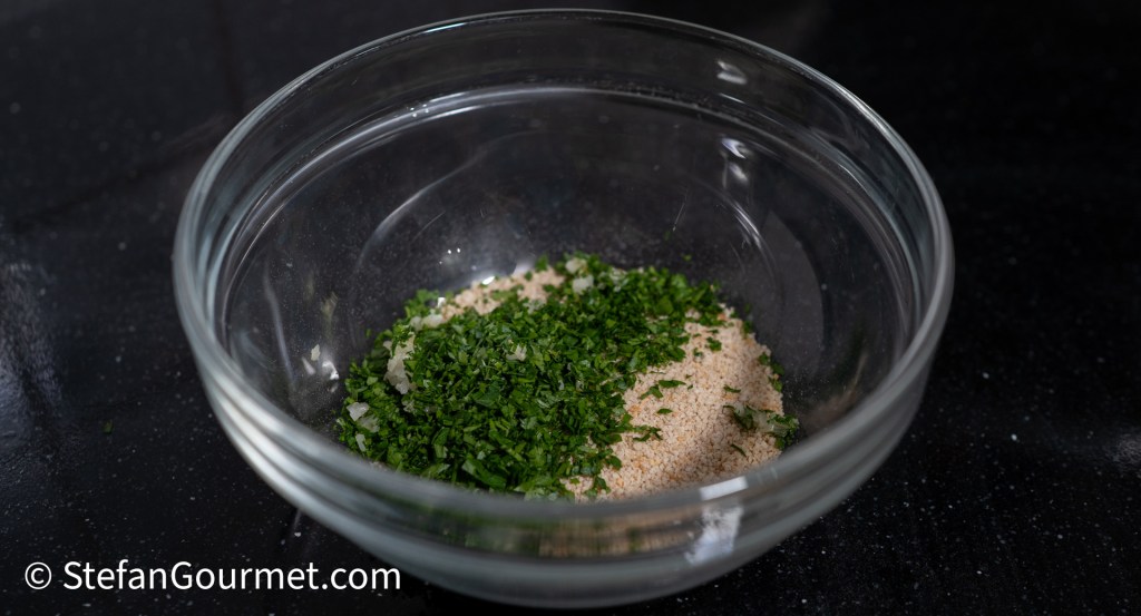 A glass bowl containing a mixture of minced fresh parsley and breadcrumbs on a dark surface.