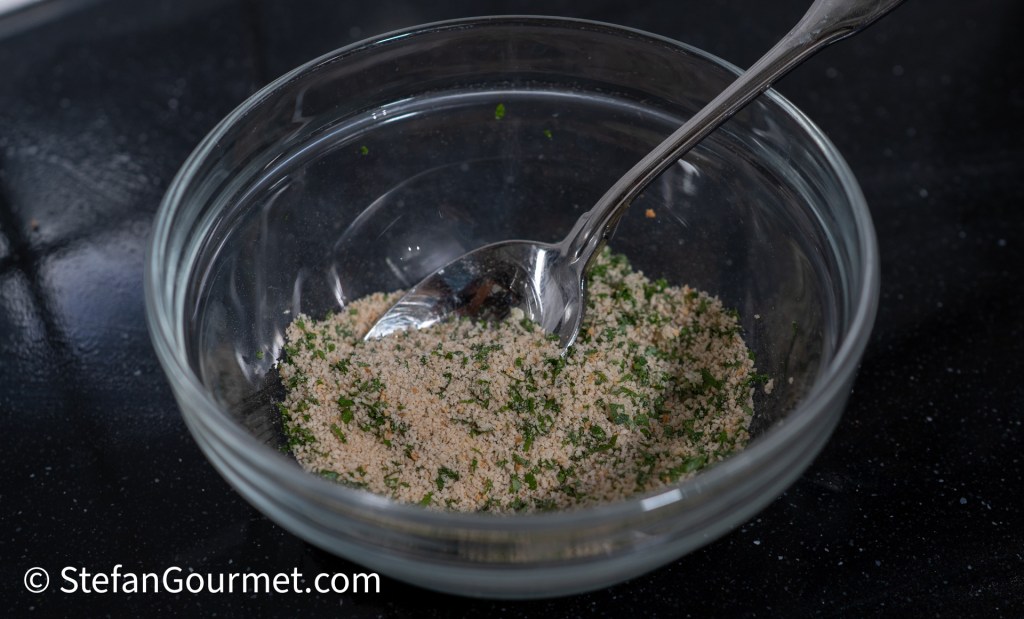 A glass bowl containing a mixture of breadcrumbs and minced parsley, with a spoon resting inside.