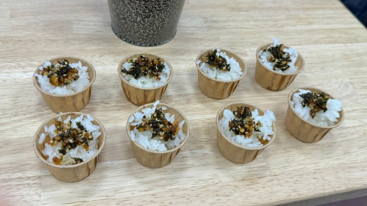 A wooden platter featuring small cups of rice topped with crispy seaweed and garnishes, arranged neatly in a casual setting.