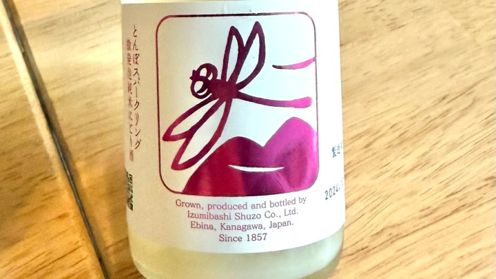 A close-up of a bottle of Izumibashi sparkling sake featuring a colorful label design with a dragonfly and stylized lips.