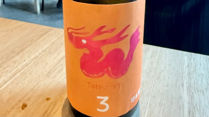 A bottle of sake labeled 'Tatsu-ism 3' featuring a vibrant pink illustration of a mythical creature on an orange background, placed on a wooden table.
